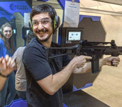 Smiling person wearing safety glasses and earmuffs holds a rifle at an indoor shooting range lane while others watch from adjacent booths.