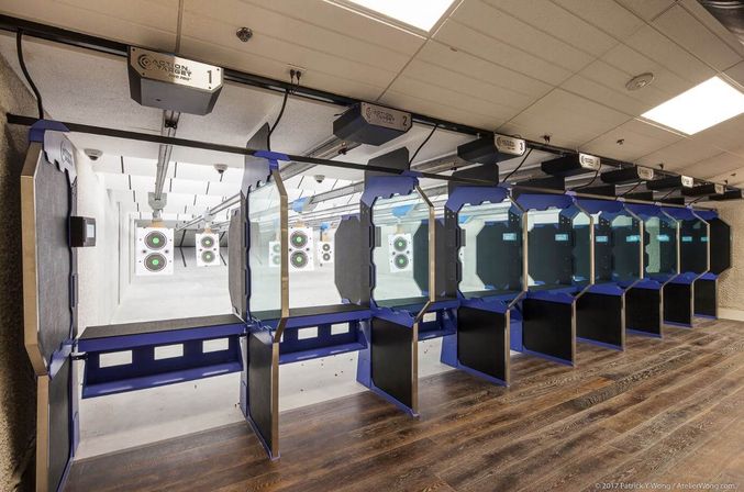Empty indoor shooting range with a row of blue glass shooting stalls, wooden floor, overhead target carriers and bullseye paper targets downrange.