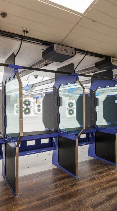 Empty indoor shooting range with a row of blue glass shooting stalls, wooden floor, overhead target carriers and bullseye paper targets downrange.