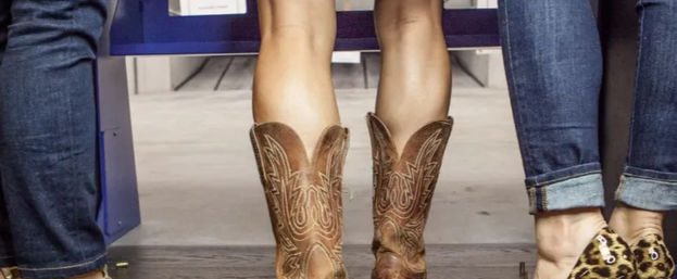 Lower legs of three people seated on a bench: center wearing brown embroidered cowboy boots, others in cuffed jeans with casual shoes including leopard-print slip-ons.