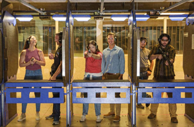 Indoor shooting range scene with people in blue-lined target lanes wearing ear protection; one person aiming a handgun downrange, another holding a rifle, and others standing nearby observing.