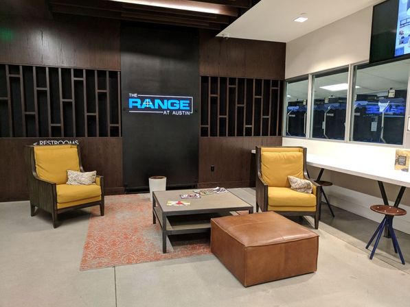 Cozy modern indoor shooting-range lounge in Austin, TX with two yellow armchairs, leather ottoman, coffee table on a patterned rug, and windows overlooking target bays.