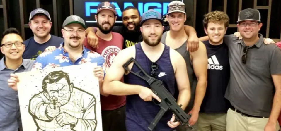 Group of smiling men at an indoor shooting range event, casually dressed with caps, one holding a black rifle and another holding a paper target showing an illustrated man aiming a gun.