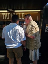 Two men in casual summer clothes at an outdoor self-pour tap station, one pointing at a touchscreen while the other watches and holds a glass, deciding a drink.