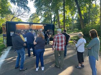 People queued at a mobile tap truck on a tree-lined suburban street, holding drinks and chatting at a casual outdoor community gathering.