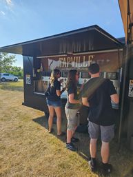 Three people using taps at a self‑pour mobile taproom trailer parked on a sunny grassy field with a blue sky overhead