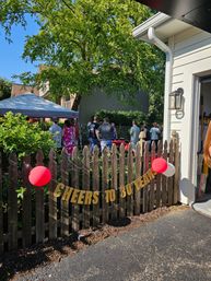 Suburban backyard party under a canopy with guests mingling, a golden "Cheers to 30 Years" banner on a wooden picket fence, red balloons, garage and trees on a sunny day