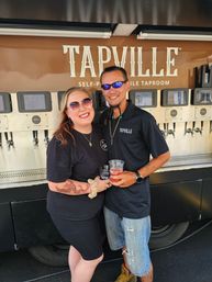 Smiling couple holding plastic beer cups in front of a self-serve mobile taproom truck with rows of beer taps; woman in sunglasses with arm tattoo and man in dark polo and sunglasses.