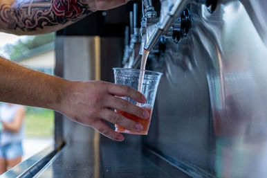 Tattooed arm tapping a stainless-steel draft, pouring a pink-hued beverage into a clear plastic cup at an outdoor event.