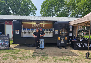 Outdoor self-serve beer trailer at a summer festival with multiple taps, two staff posing in front, a service tent and leafy park background.