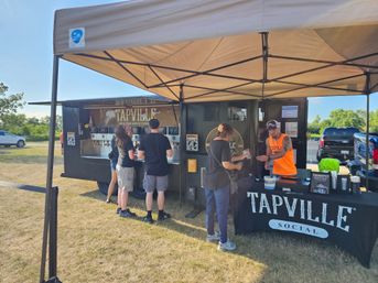 Sunny outdoor self-serve tap trailer under a beige canopy, customers queuing on a grassy field at a casual summer event
