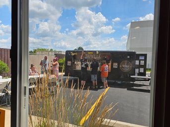 Sunny outdoor patio scene: people queuing at a black mobile beer tap trailer in a parking lot, folding tables and chairs, ornamental grasses in foreground and blue sky with fluffy clouds.