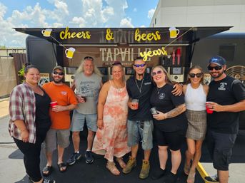Eight people smiling and posing in front of a mobile beer tap trailer at an outdoor summer parking-lot gathering, holding cups beneath a festive "cheers & beers" banner.