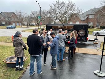 Outdoor neighborhood gathering in a suburban driveway — people socializing with drinks around a mobile tap trailer and fire pit on an overcast day.