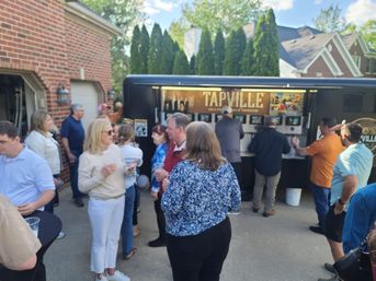 Group of people gathered around a black mobile tap trailer in a suburban driveway, chatting and holding plastic cups at a casual outdoor party.