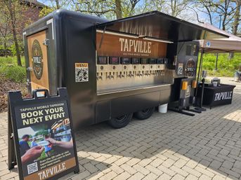Self-pour mobile taproom trailer with row of stainless beer taps, A-frame “Book your next event” sign and QR code, canopy and service table on brick-paved path in a leafy park.