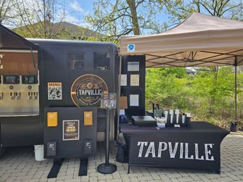 Outdoor mobile taproom and canopy with self-serve beer taps, cups, QR-code menu and express checkout on a paved plaza beside leafy green trees.