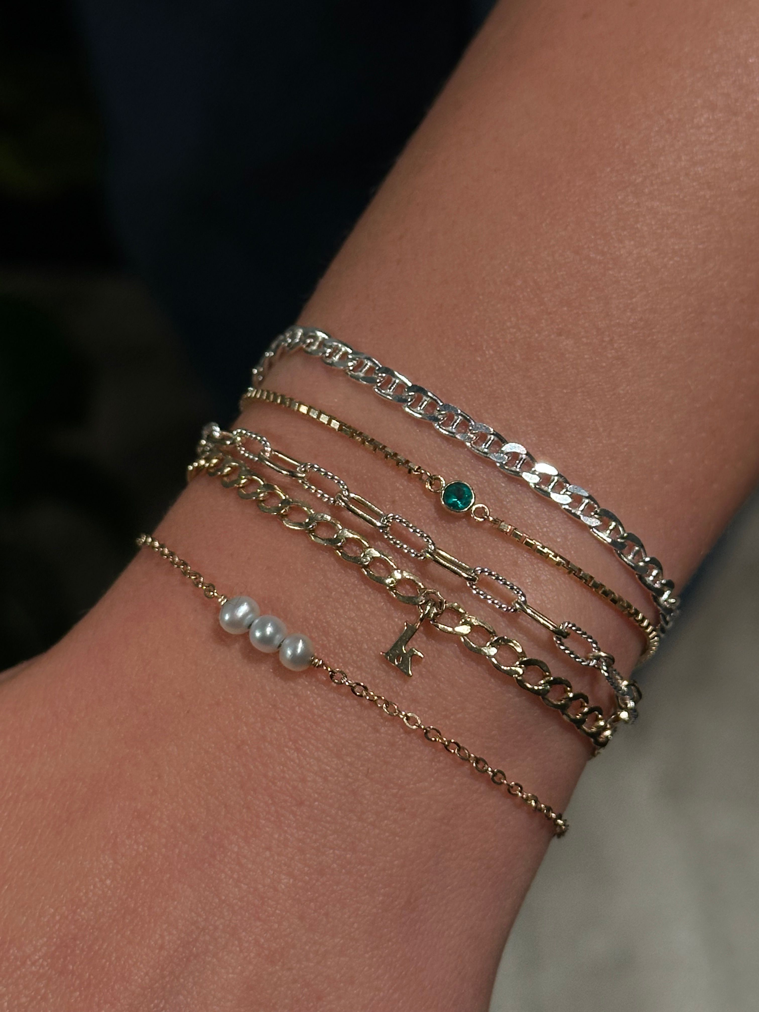 Close-up of a wrist wearing stacked delicate gold and silver chain bracelets with three tiny pearls, a small green gemstone and a dainty letter charm — layered minimalist jewelry, perfect for everyday style.