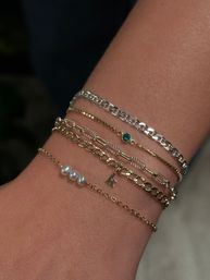 Close-up of a wrist wearing stacked delicate gold and silver chain bracelets with three tiny pearls, a small green gemstone and a dainty letter charm — layered minimalist jewelry, perfect for everyday style.