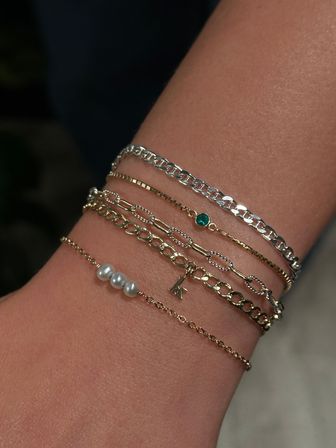 Close-up of a wrist wearing stacked delicate gold and silver chain bracelets with three tiny pearls, a small green gemstone and a dainty letter charm — layered minimalist jewelry, perfect for everyday style.