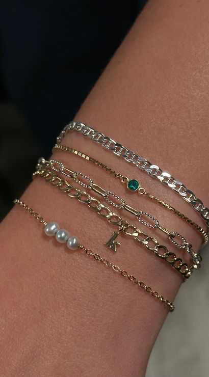 Close-up of a wrist wearing stacked delicate gold and silver chain bracelets with three tiny pearls, a small green gemstone and a dainty letter charm — layered minimalist jewelry, perfect for everyday style.