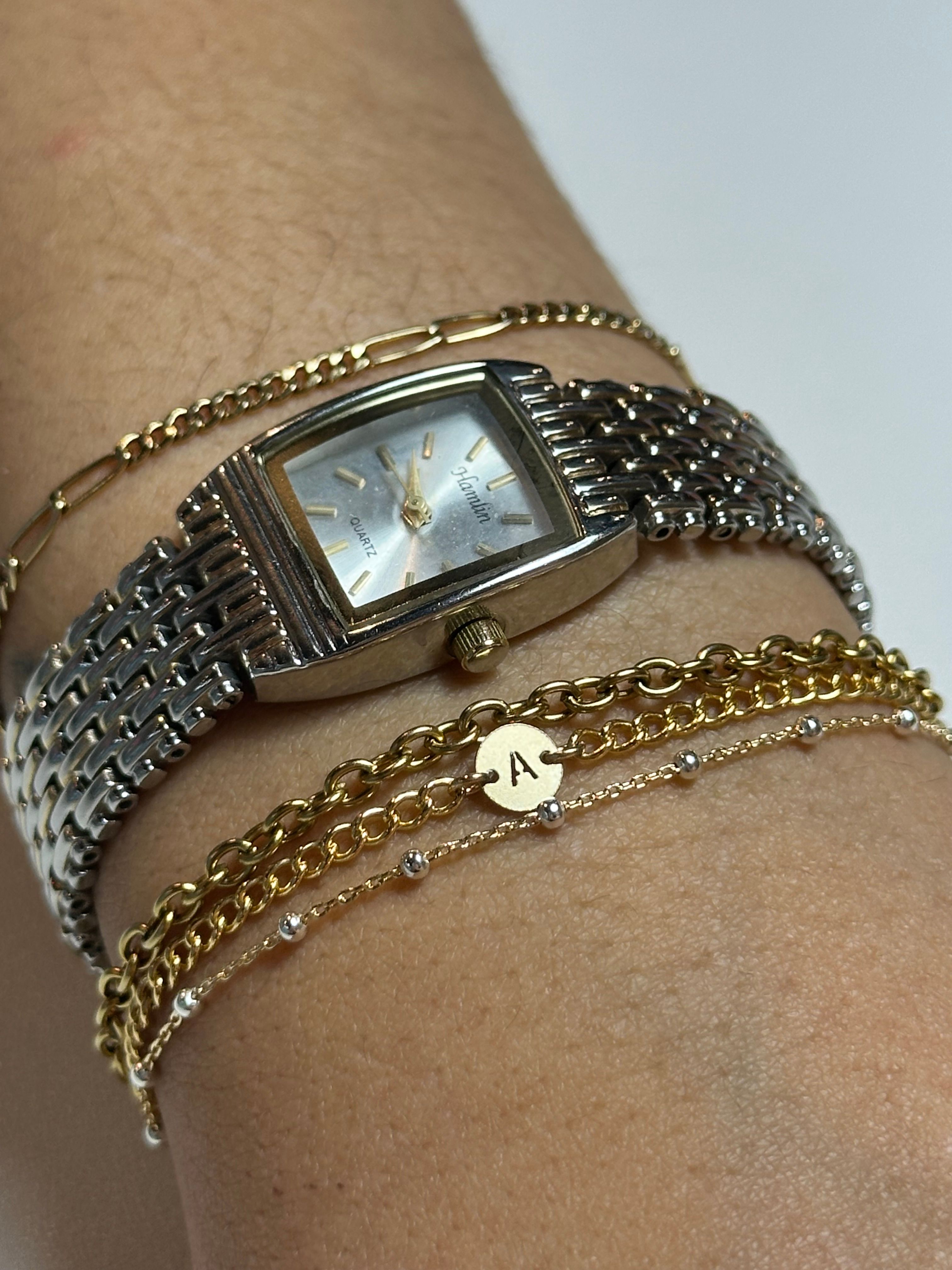 Close-up of a wrist wearing a silver rectangular quartz watch with woven metal band and stacked gold chain bracelets, featuring a small round disc charm engraved with the letter A.