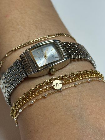 Close-up of a wrist wearing a silver rectangular quartz watch with woven metal band and stacked gold chain bracelets, featuring a small round disc charm engraved with the letter A.