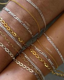 Close-up of two wrists adorned with stacked mixed-metal chain bracelets — gold and silver Figaro, curb and box-link styles layered on sunlit skin