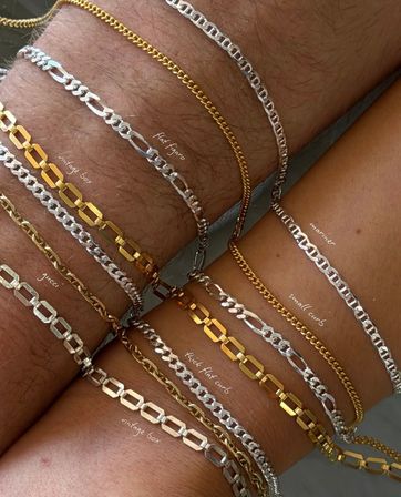 Close-up of two wrists adorned with stacked mixed-metal chain bracelets — gold and silver Figaro, curb and box-link styles layered on sunlit skin