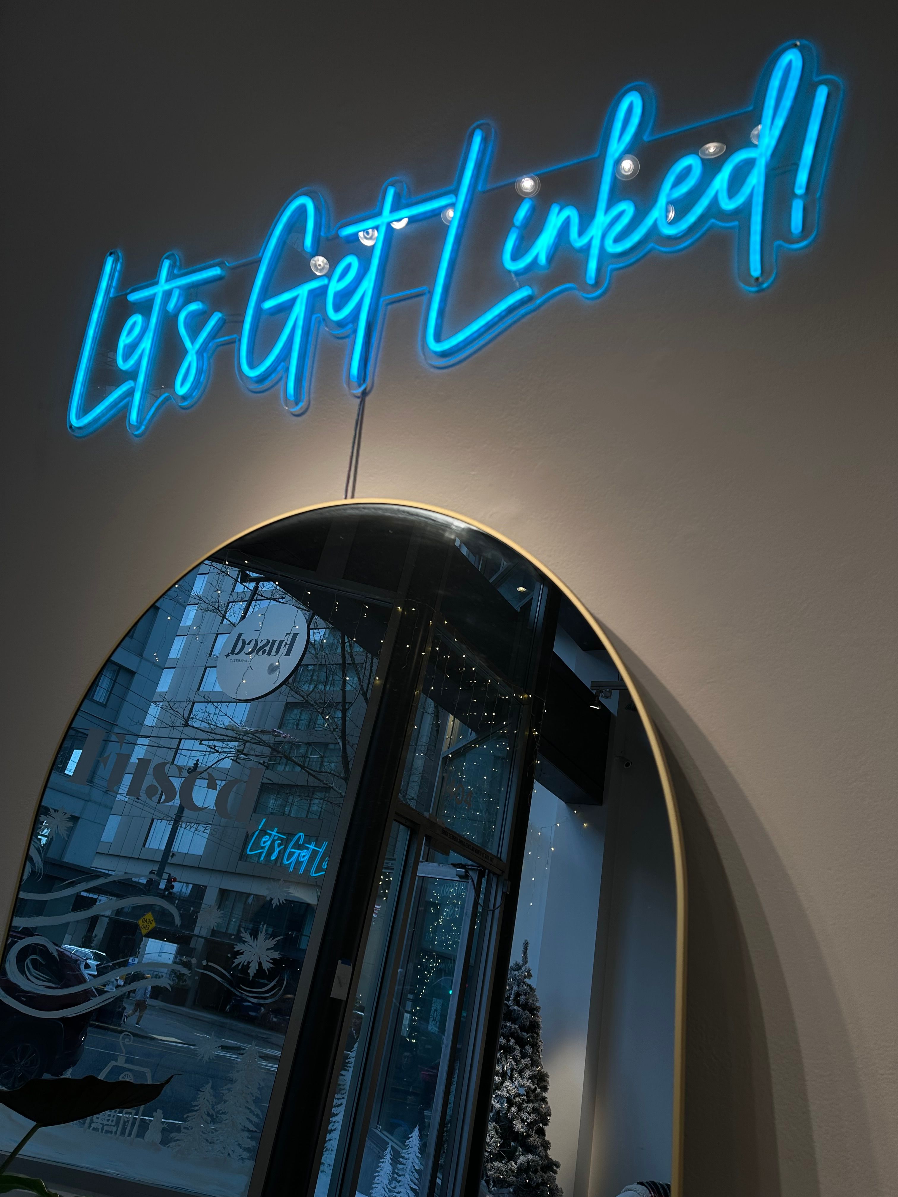 Playful blue neon sign reading "Let's Get Linked!" above a round gold-framed mirror reflecting a downtown storefront, snowy holiday tree, string lights and city street scene.