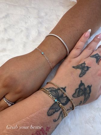 Close-up of two wrists on white faux fur showing stacked bracelets and friendship chains — a silver snake bangle and delicate silver chain with tiny blue gem on one wrist, layered gold chains with a small blue stone on a butterfly-tattooed wrist for a playful 'grab your bestie' jewelry vibe.