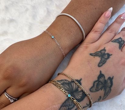 Close-up of two wrists on white faux fur showing stacked bracelets and friendship chains — a silver snake bangle and delicate silver chain with tiny blue gem on one wrist, layered gold chains with a small blue stone on a butterfly-tattooed wrist for a playful 'grab your bestie' jewelry vibe.