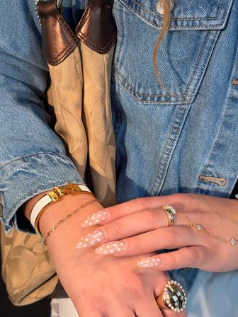 Close-up of hands with long stiletto white polka-dot manicure, stacked gold rings and delicate chain bracelets, styled against a blue denim jacket and beige patterned bag strap — chic street-style accessory shot.