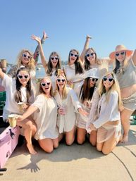 Smiling group of women in white outfits and heart-shaped sunglasses celebrating on a sunny boat near a harbor, holding cups for a playful bachelorette-style party vibe