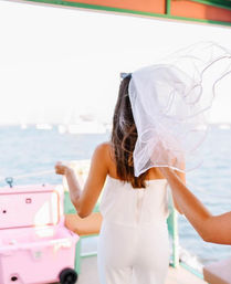 Bride-to-be in a white outfit on a sunny boat deck, veil blowing in the sea breeze with a pink cooler and ocean in the background.