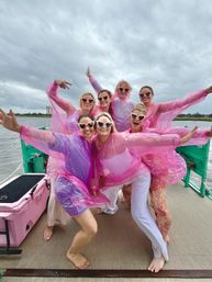 Seven women on a boat deck wearing pink translucent rain ponchos and heart-shaped sunglasses, posing joyfully against a cloudy waterfront sky.