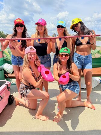 Six friends on a sunny pontoon boat wearing colorful caps and swimsuits, holding a pink shot-ski with cup holders and two pink flamingo drink holders, lake and cloudy sky in the background.