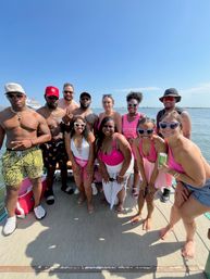 Smiling group of friends on a pontoon boat under a clear blue sky, many wearing pink swimwear and heart-shaped sunglasses, posing for a summer waterfront boat party with a cruise ship and shoreline visible in the background.
