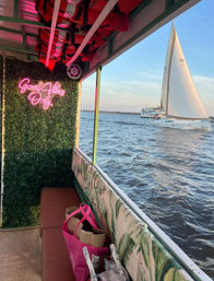 View from a decorated waterfront boat interior with neon “Good Vibes Only” sign, tropical-pattern bench, pink tote bags and life jackets overhead, looking out at a white sailboat on open water at sunset.