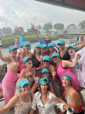 Cheerful group of women in matching teal caps and colorful summer outfits posing and cheering on a boat at a marshy waterfront dock in light rain, many holding drinks.