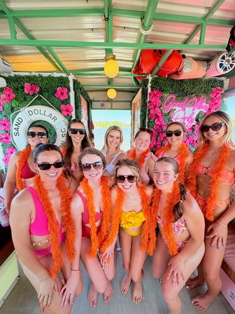 Ten friends in colorful swimsuits and sunglasses wearing bright orange feather boas, smiling and posing on a decorated boat with pink flower walls, a life preserver, neon “Good Vibes” sign, and a lake visible through the doorway — summer girls‑trip boat party vibe.