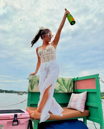Stylish woman in a white lace top and sarong stands on a colorful boat bench, raising a champagne bottle over calm coastal waters, summer boat party vibe.