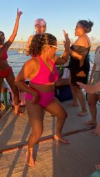 Group of women dancing on a boat at golden hour, central woman in a hot-pink bikini striking a playful pose with water and a bridge in the background.
