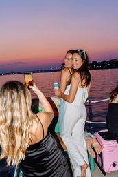 Two friends posing for a selfie on a boat at a pink‑purple sunset over a calm harbor, smiling and holding canned drinks.