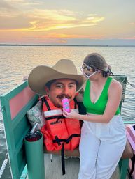 Woman in a green crop top on a colorful pontoon boat at sunset holding a pink can cooler up to a giant novelty cowboy-face cutout wearing a hat and life jacket, calm water and orange sky in the background.