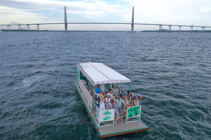 Happy crowd on a colorful covered party pontoon boat cruising a coastal bay, with a long cable-stayed bridge on the horizon and a cloudy sky.