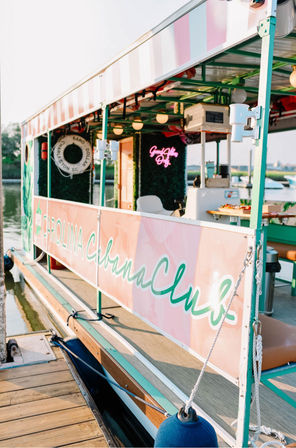 Sunlit pink-and-teal party boat moored at a wooden dock with a striped canopy, life preserver, neon sign, seating and a small snack counter, calm marina in the background.