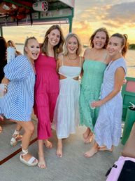 Five friends in colorful summer dresses posing barefoot on a boat deck during a coastal sunset cruise, smiling against a golden sky and calm water.