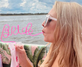 Profile of a person wearing white heart-shaped sunglasses sipping from a neon-pink curly straw that spells "bride" aboard a boat with tropical-print cushions and a cloudy waterfront backdrop.
