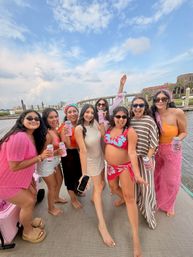 Eight friends smiling and posing on a boat deck at a sunny waterfront marina, wearing colorful swimsuits and summer outfits and holding canned drinks with a bridge and shoreline in the background.